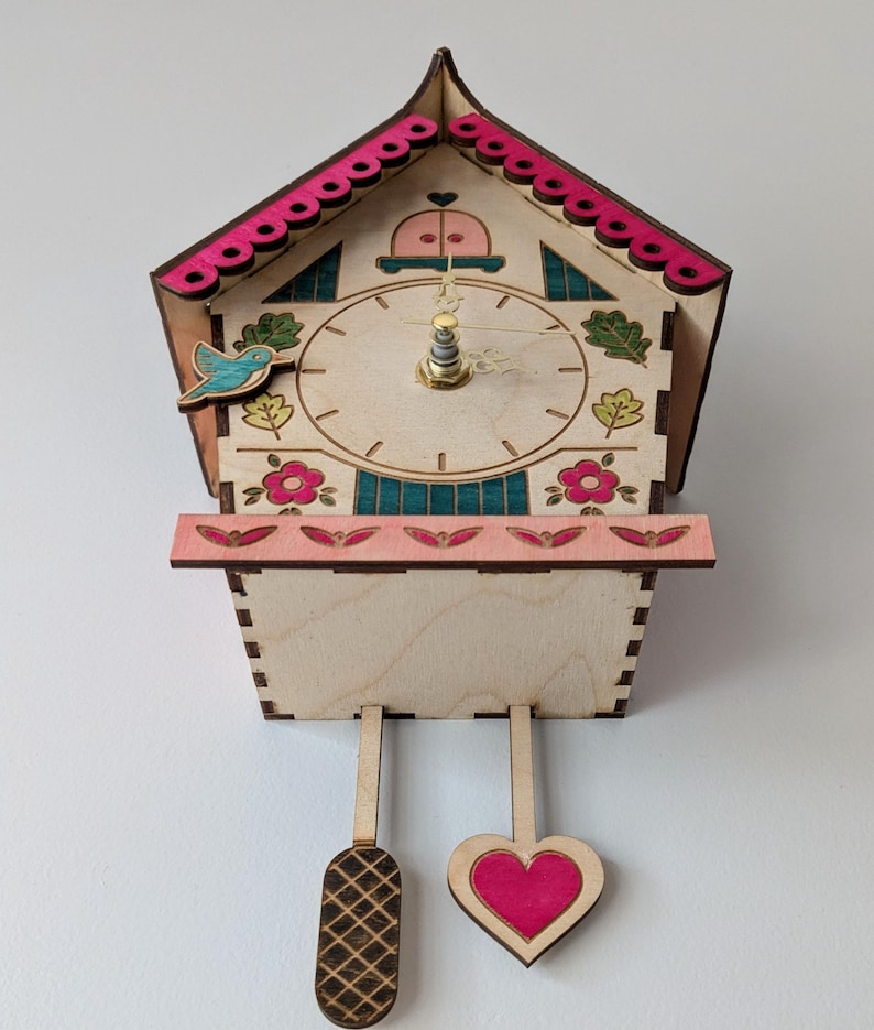 May include: A wooden cuckoo clock with a pink and white painted design. The clock face has a blue bird, pink flowers, and green leaves. The clock has a heart-shaped pendulum and a rectangular pendulum.