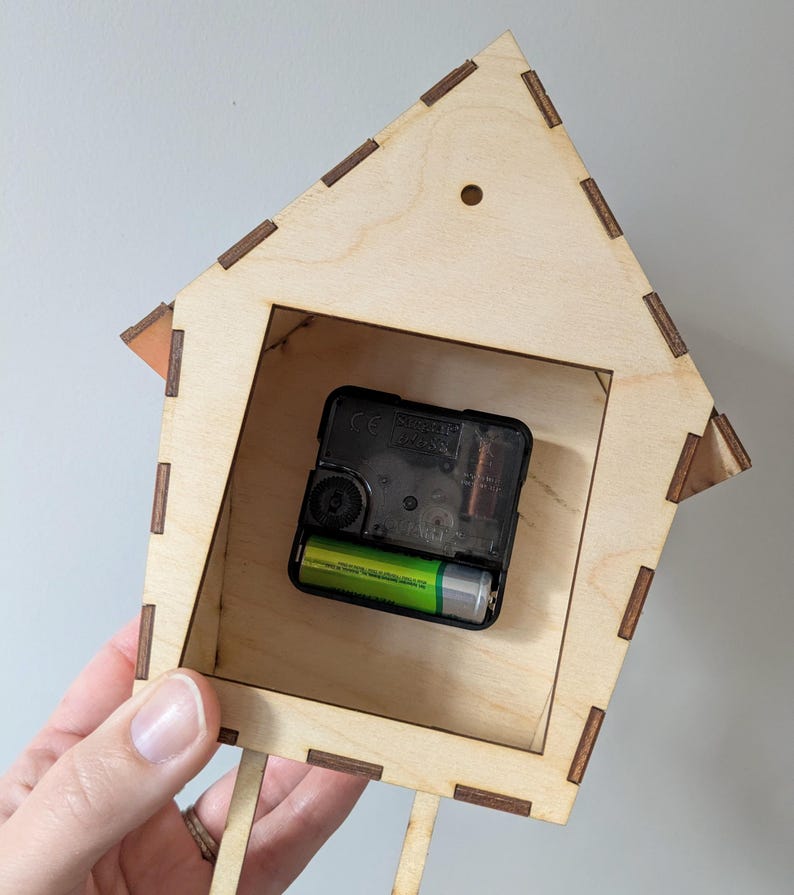 May include: A wooden house-shaped clock with a black clock mechanism and a green battery inside. The clock is made of light wood with dark brown accents.