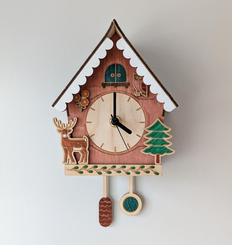 May include: A wooden cuckoo clock shaped like a house with a brown and white exterior. The clock face is surrounded by a deer, a tree, and an owl. The clock has a pendulum with a pine cone and a green circle.