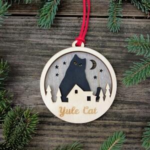 May include: Wooden Christmas ornament with a black cat silhouette, stars, and a crescent moon. The ornament has the words "Yule Cat" engraved on it. It is hanging from a red string, with evergreen branches in the background.