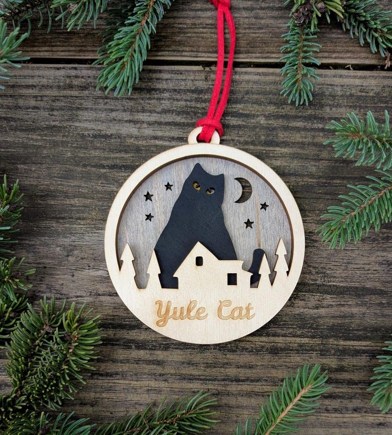 May include: Wooden Christmas ornament with a black cat silhouette, stars, and a crescent moon. The ornament has the words "Yule Cat" engraved on it. It is hanging from a red string, with evergreen branches in the background.