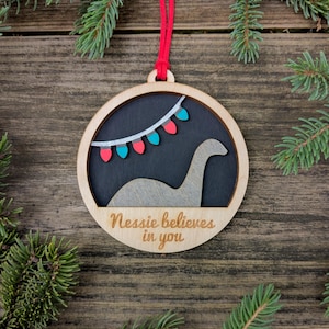 May include: Wooden Christmas ornament with a circular design. The ornament features a silhouette of the Loch Ness Monster, a string of colorful lights, and the text "Nessie believes in you". A red string is attached for hanging.
