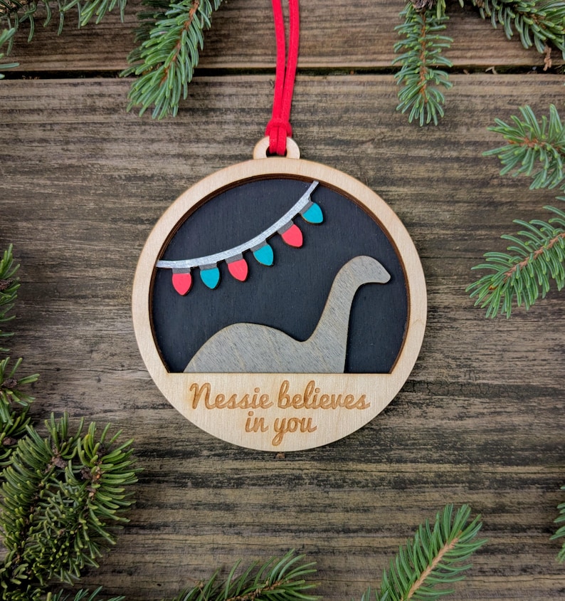 May include: Wooden Christmas ornament with a circular design. The ornament features a silhouette of the Loch Ness Monster, a string of colorful lights, and the text "Nessie believes in you". A red string is attached for hanging.
