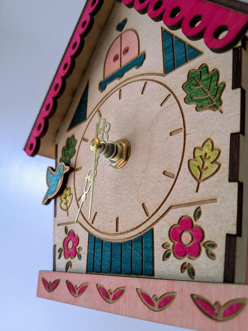 May include: A wooden cuckoo clock shaped like a house with a pink roof and blue and green accents. The clock face has a gold-colored hour and minute hand. The clock is decorated with flowers, leaves, and a bird.