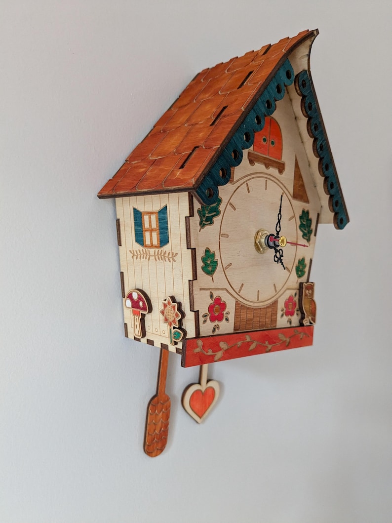 May include: A wooden cuckoo clock shaped like a house with a red roof, green trim, and a white face. The clock has a pendulum and a small owl on the side. The clock face has Roman numerals and a red second hand.
