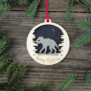 May include: Wooden Christmas ornament with a cryptid design. The ornament features a light-colored wooden frame with a cutout of a forest silhouette. Inside, a gray cryptid walks under a night sky with stars and a crescent moon. The text "Merry Cryptmas" is engraved.