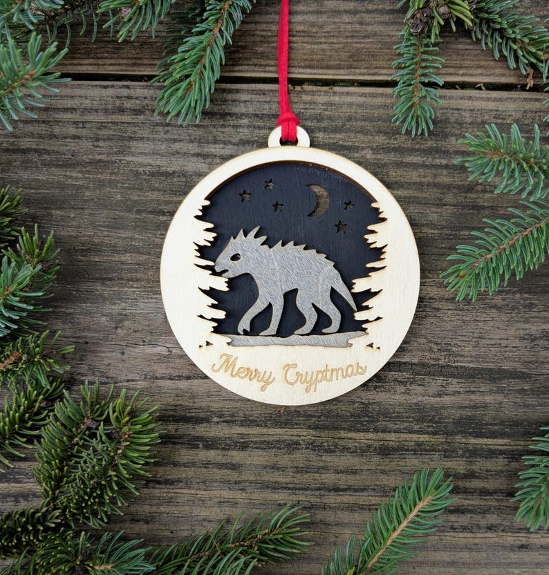 May include: Wooden Christmas ornament with a cryptid design. The ornament features a light-colored wooden frame with a cutout of a forest silhouette. Inside, a gray cryptid walks under a night sky with stars and a crescent moon. The text "Merry Cryptmas" is engraved.
