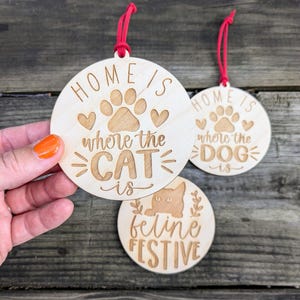 May include: Three wooden ornaments with red string hangers. One ornament says "Home is where the CAT is" with a paw print and hearts. Another says "Home is where the DOG is". The third says "feline FESTIVE" with a cat illustration.