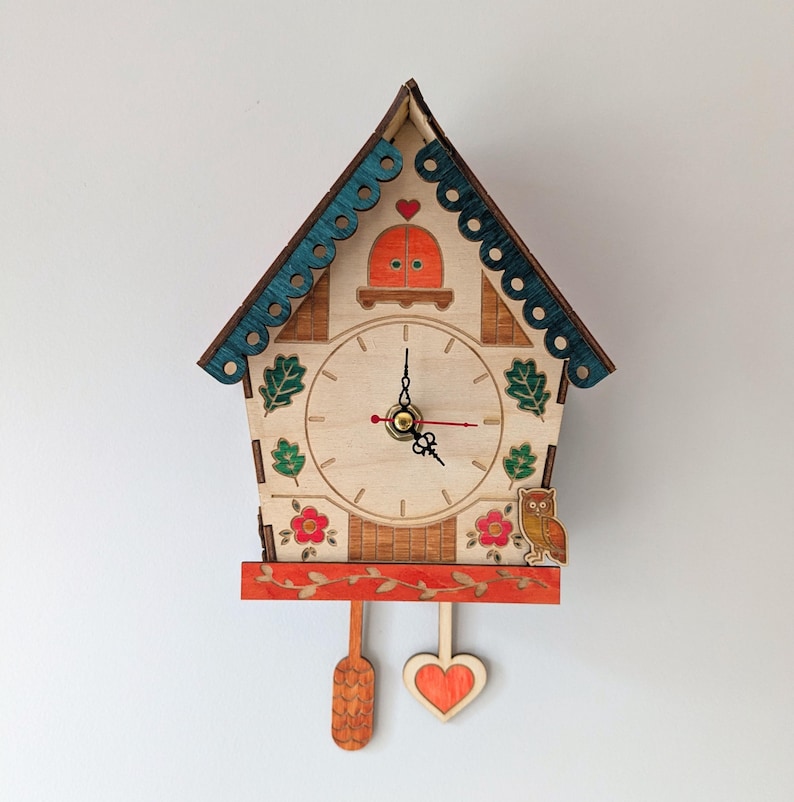 May include: A wooden cuckoo clock shaped like a house with a red heart, green leaves, and an owl. The clock face has black hands and Roman numerals.