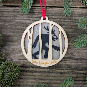 May include: Wooden Christmas ornament with a black silhouette of a creature with antlers, framed by birch trees. The ornament has the words "Live Laugh Lurk" and is suspended by a red string. The background is a wooden surface with pine needles.