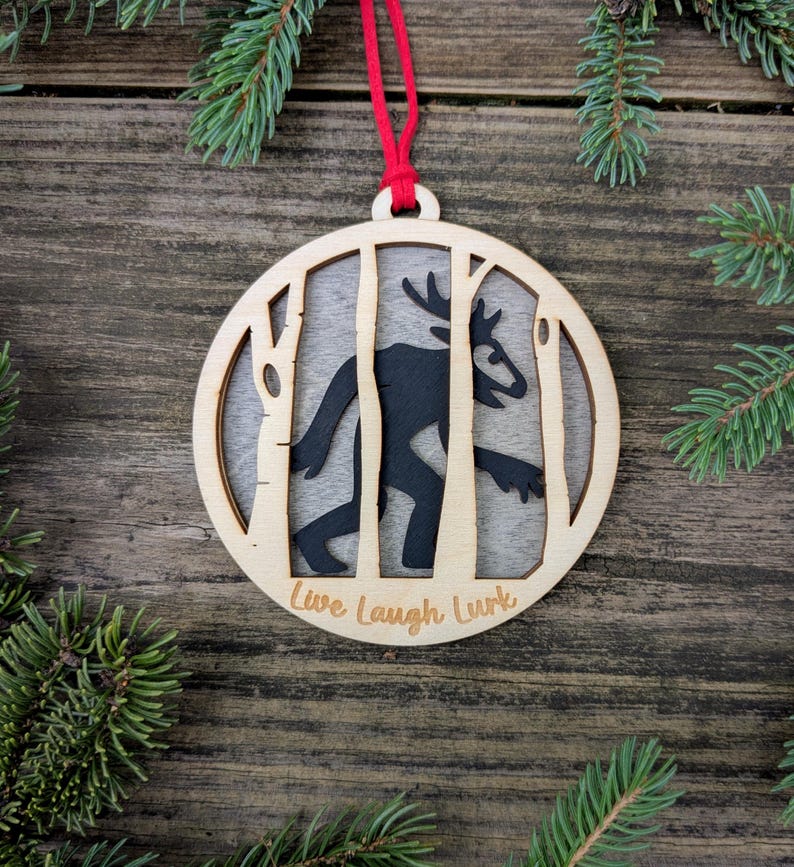 May include: Wooden Christmas ornament with a black silhouette of a creature with antlers, framed by birch trees. The ornament has the words "Live Laugh Lurk" and is suspended by a red string. The background is a wooden surface with pine needles.