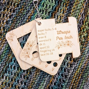 May include: Wooden gauge with holes for measuring yarn wraps per inch. The gauge is labelled with different yarn weights, including super bulky, bulky, aran, worsted, dk, sport, fingering, and lace. The gauge is also labelled with "Wraps Per Inch" and has a small floral design.