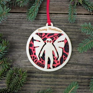May include: Wooden Christmas ornament with a layered design featuring a figure with wings, set against a red and black background. The ornament has the words "Merry Mothmas" engraved on the top and is suspended by a red cord.