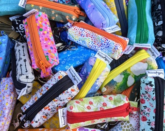 May include: A collection of small, colorful zippered pouches with various patterns, including floral, polka dots, and animal prints. Each pouch has a contrasting zipper and a small tag. The pouches are made of fabric and have a rectangular shape.