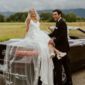 K&ouml;nnte beinhalten: Eine Braut in einem wei&szlig;en Satin-Hochzeitskleid und Schleier sitzt auf der Motorhaube eines schwarzen Cabriolets. Ein Mann in einem schwarzen Smoking steht daneben und l&auml;chelt. Der Hintergrund zeigt ein Feld und Berge.