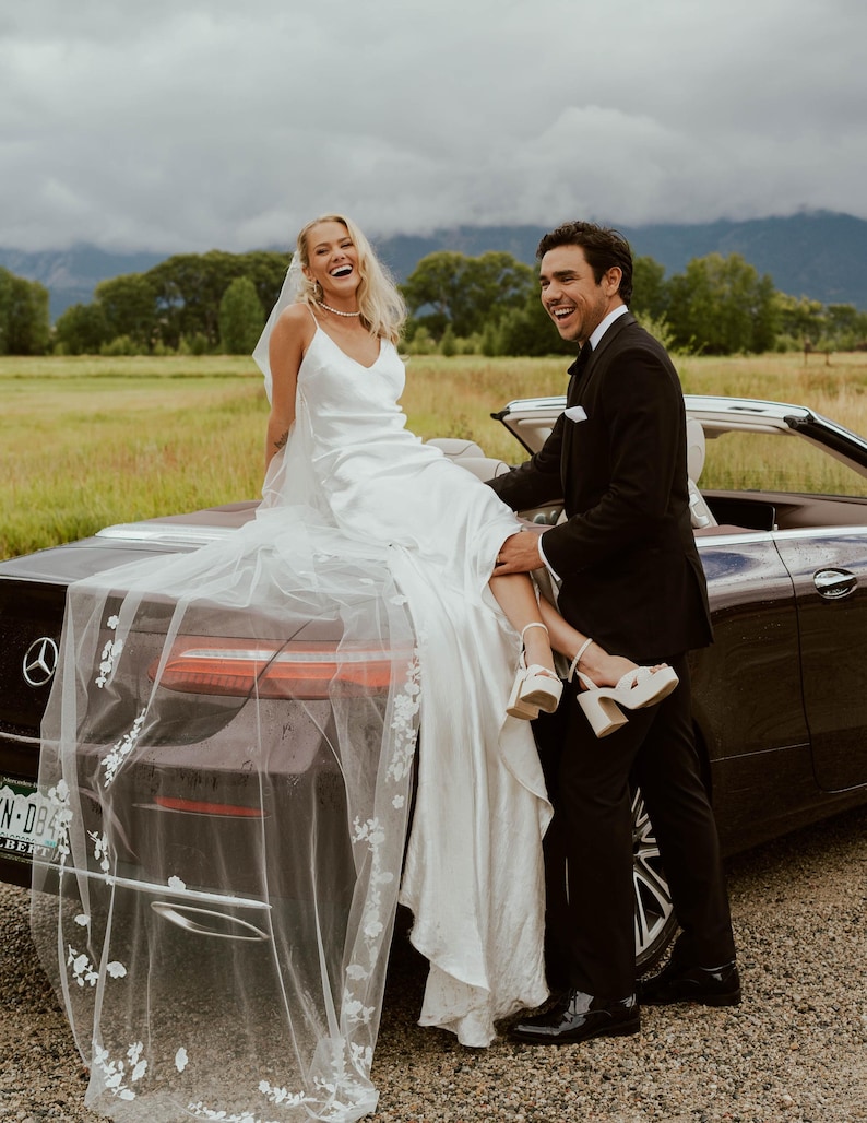 K&ouml;nnte beinhalten: Eine Braut in einem wei&szlig;en Satin-Hochzeitskleid und Schleier sitzt auf der Motorhaube eines schwarzen Cabriolets. Ein Mann in einem schwarzen Smoking steht daneben und l&auml;chelt. Der Hintergrund zeigt ein Feld und Berge.