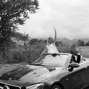 K&ouml;nnte beinhalten: Schwarzes Cabrio mit einer Frau in einem wei&szlig;en Kleid, die von oben winkt. Ein Mann im Anzug winkt vom Fahrersitz aus. Das Auto parkt auf einer Stra&szlig;e mit B&auml;umen und Bergen im Hintergrund.