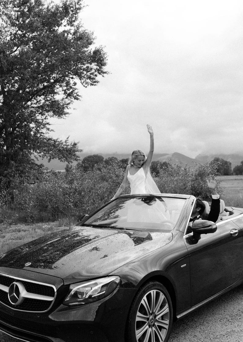 K&ouml;nnte beinhalten: Schwarzes Cabrio mit einer Frau in einem wei&szlig;en Kleid, die von oben winkt. Ein Mann im Anzug winkt vom Fahrersitz aus. Das Auto parkt auf einer Stra&szlig;e mit B&auml;umen und Bergen im Hintergrund.