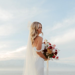 K&ouml;nnte beinhalten: Eine Braut in einem wei&szlig;en schulterfreien Brautkleid h&auml;lt einen Strau&szlig; roter, rosa und gelber Blumen. Sie tr&auml;gt einen Schleier und hat blondes Haar. Der Hintergrund zeigt einen hellblauen Himmel und ein Gew&auml;sser.
