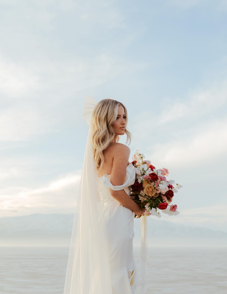 K&ouml;nnte beinhalten: Eine Braut in einem wei&szlig;en schulterfreien Brautkleid h&auml;lt einen Strau&szlig; roter, rosa und gelber Blumen. Sie tr&auml;gt einen Schleier und hat blondes Haar. Der Hintergrund zeigt einen hellblauen Himmel und ein Gew&auml;sser.