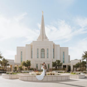K&ouml;nnte beinhalten: Ein Paar in Hochzeitskleidung steht vor einem gro&szlig;en, wei&szlig;en Geb&auml;ude mit einem hohen Turm und einer goldenen Statue. Palmen und Gr&uuml;n umgeben das Geb&auml;ude. Der Himmel ist blau mit Wolken. Die Szene deutet auf eine Hochzeit hin.