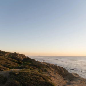 K&ouml;nnte beinhalten: Eine malerische K&uuml;stenlandschaft bei Sonnenuntergang. Ein Paar steht auf einer Klippe mit Blick auf das Meer. Der Himmel wechselt von Blau zu Orange und wirft einen warmen Schein auf die Szene. Der Hang ist mit gr&uuml;ner und brauner Vegetation bedeckt.