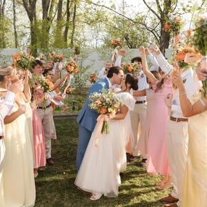 K&ouml;nnte beinhalten: Eine Hochzeitsfeier mit einem sich k&uuml;ssenden Brautpaar, umgeben von G&auml;sten, die Blumenstr&auml;u&szlig;e halten. Die Braut tr&auml;gt ein wei&szlig;es Kleid, der Br&auml;utigam einen blauen Anzug. G&auml;ste tragen Pastellfarben, im Freien.