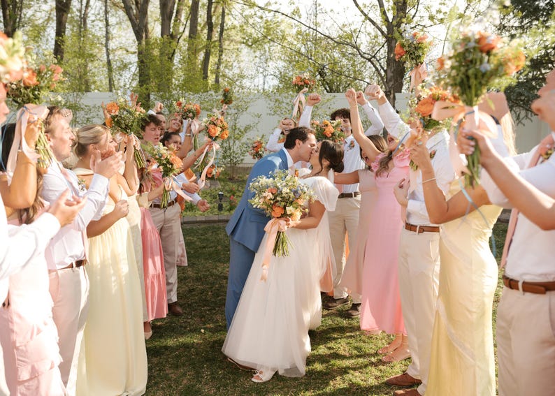 K&ouml;nnte beinhalten: Eine Hochzeitsfeier mit einem sich k&uuml;ssenden Brautpaar, umgeben von G&auml;sten, die Blumenstr&auml;u&szlig;e halten. Die Braut tr&auml;gt ein wei&szlig;es Kleid, der Br&auml;utigam einen blauen Anzug. G&auml;ste tragen Pastellfarben, im Freien.
