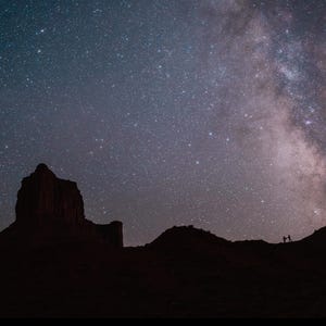K&ouml;nnte beinhalten: Nachtlandschaft mit einer silhouetten Bergkette und einem lebendigen Sternenhimmel. Die Milchstra&szlig;e ist sichtbar, mit einem hell leuchtenden Sternenband. Zwei Figuren stehen auf einem fernen Grat und geben der Szene Ma&szlig;stab.