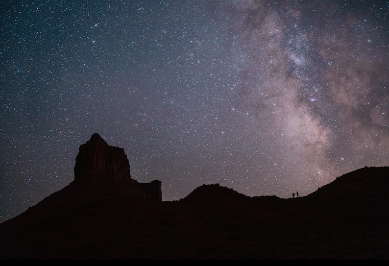 K&ouml;nnte beinhalten: Nachtlandschaft mit einer silhouetten Bergkette und einem lebendigen Sternenhimmel. Die Milchstra&szlig;e ist sichtbar, mit einem hell leuchtenden Sternenband. Zwei Figuren stehen auf einem fernen Grat und geben der Szene Ma&szlig;stab.