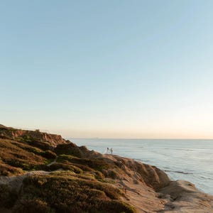 K&ouml;nnte beinhalten: Eine malerische K&uuml;stenlandschaft mit einer Klippe, die mit brauner Vegetation bewachsen ist und auf das Meer blickt. Der Himmel wechselt von hellblau zu orange, was auf einen Sonnenuntergang hindeutet. Zwei Figuren stehen auf der Klippe.