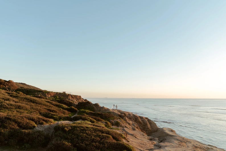 K&ouml;nnte beinhalten: Eine malerische K&uuml;stenlandschaft mit einer Klippe, die mit brauner Vegetation bewachsen ist und auf das Meer blickt. Der Himmel wechselt von hellblau zu orange, was auf einen Sonnenuntergang hindeutet. Zwei Figuren stehen auf der Klippe.