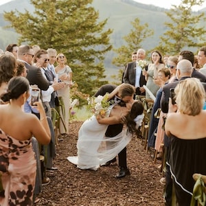 K&ouml;nnte beinhalten: Hochzeitsszene: Braut und Br&auml;utigam k&uuml;ssen sich, w&auml;hrend die G&auml;ste applaudieren. Die Braut tr&auml;gt ein wei&szlig;es Kleid und einen Blumenstrau&szlig;. Der Br&auml;utigam tr&auml;gt einen schwarzen Anzug. Die Zeremonie findet im Freien mit Bergkulisse statt.