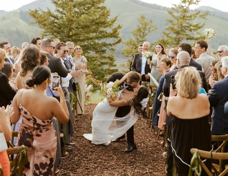 K&ouml;nnte beinhalten: Hochzeitsszene: Braut und Br&auml;utigam k&uuml;ssen sich, w&auml;hrend die G&auml;ste applaudieren. Die Braut tr&auml;gt ein wei&szlig;es Kleid und einen Blumenstrau&szlig;. Der Br&auml;utigam tr&auml;gt einen schwarzen Anzug. Die Zeremonie findet im Freien mit Bergkulisse statt.