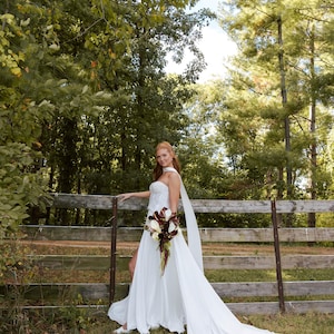 K&ouml;nnte beinhalten: Wei&szlig;es Hochzeitskleid mit langer Schleppe und tr&auml;gerlosem Oberteil. Das Kleid wird mit einem Strau&szlig; wei&szlig;er und burgunderfarbener Blumen kombiniert. Die Braut steht neben einem Holzzaun in einer nat&uuml;rlichen Umgebung im Freien.