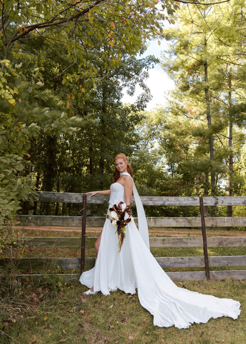 K&ouml;nnte beinhalten: Wei&szlig;es Hochzeitskleid mit langer Schleppe und tr&auml;gerlosem Oberteil. Das Kleid wird mit einem Strau&szlig; wei&szlig;er und burgunderfarbener Blumen kombiniert. Die Braut steht neben einem Holzzaun in einer nat&uuml;rlichen Umgebung im Freien.
