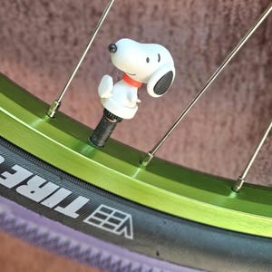 May include: A Snoopy valve stem cap is attached to a green bicycle wheel. The white Snoopy figure has a black ear and a red collar. The tire is black with white lettering.