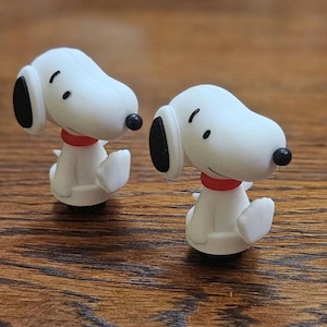 May include: Two small Snoopy figurines. Each figurine is white with black ears, a black nose, and a red collar. The figurines are sitting on a wooden surface.