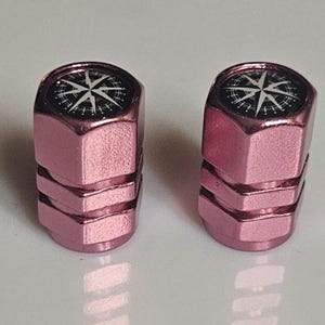 May include: Two pink metal tire valve stem caps. Each cap has a black and white compass rose design on top. The caps are hexagonal in shape and have a metallic finish. These caps are a decorative accessory for vehicles.