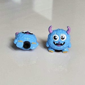 以下が含まれることがあります： 大きな目、角、笑顔の口を持つ、2つの小さな青いモンスターの置物。1つは直立しており、もう1つは仰向けになっています。モンスターは黄色い足と紫色の角を持っています。これらのおもちゃはおそらく子供向けです。