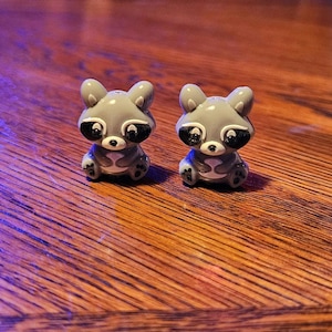 May include: Two gray raccoon figurines with black eye masks and white accents sit on a wooden surface. The raccoons have small, rounded ears and are in a sitting position. The figurines are detailed and have a cartoonish appearance.
