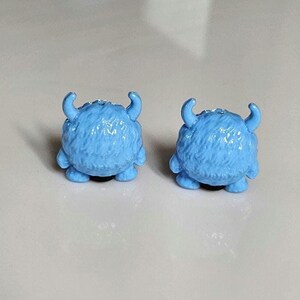 以下が含まれることがあります： 水色のモンスター型のイヤリングのペア。各イヤリングは、毛皮の質感のある丸い体、小さな腕、湾曲した角が特徴です。イヤリングは黒いポストにセットされています。