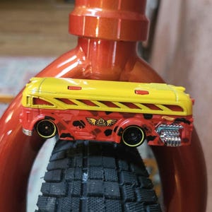 May include: A red and yellow toy bus with black wheels, set against a red metal frame and a black tire. The bus features a camouflage design and the Hot Wheels logo.
