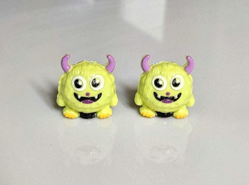 以下が含まれることがあります： モンスター型のピアス。各ピアスは黄色の体、紫の角、白黒の目があります。ピアスは笑顔の口とオレンジ色の足を持っています。これらは楽しいアクセサリーです。