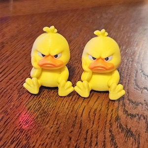 May include: Two yellow duck figurines with an angry expression. The ducks have orange beaks and feet, and are sitting down. The figurines are on a wooden surface.