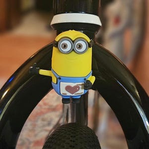 May include: A yellow Minion character with blue overalls and a heart on the front is attached to a black bicycle frame.