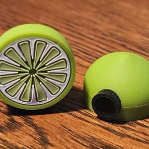 May include: Two lime-shaped bottle stoppers. One stopper is a lime slice with a silver rim and green segments. The other is a solid green dome with a black opening. Both are on a wooden surface.