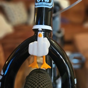 May include: A white duck-shaped bicycle accessory with orange feet and a yellow beak is attached to the front tire of a black bicycle. The bicycle frame has the text "OTW OFF THE WALL" on the frame.