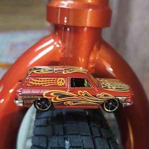 May include: A miniature red and gold station wagon toy car with a peace symbol on the side. The car has black wheels and is decorated with flame and swirl designs. The car is resting on a black tire.