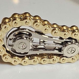 Fidget: A gold-colored bicycle chain link design with silver-colored mechanical components. The chain is arranged in a loop around the silver-colored parts. The object is on a white surface.
